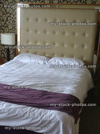 Stock image of kingsize double bed, large beige studded leather headboard, white sheets, bedside lamps
