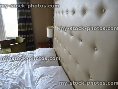 Stock image of kingsize double bed, large beige studded leather headboard, white sheets, bedside lamp