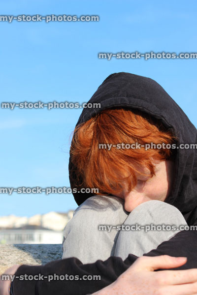 Stock image of red hair teenage boy wearing black hoodie, youth curled up, foetus position