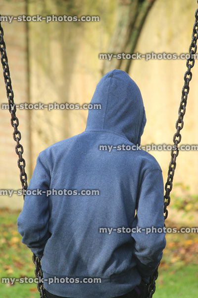 Stock image of lonely teenage boy / youth wearing hoodie / hood, sitting playground swing