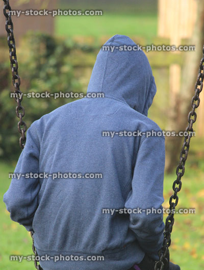 Stock image of lonely teenage boy / youth wearing hoodie / hood, sitting playground swing