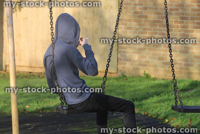 Stock image of teenage boy smoking underage / youth wearing hoodie / hood, playground swing