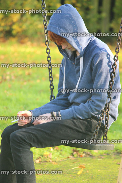 Stock image of teenage boy smoking underage / youth wearing hoodie / hood, playground swing