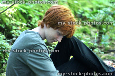 Stock image of teenage boy sitting in woodland / forest, daydreaming, looking at floor