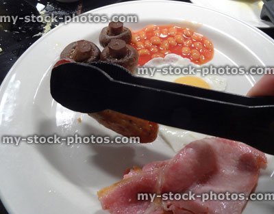 Stock image of full English fried breakfast served with tongs, sausages, bacon, fried egg, fried mushrooms, baked beans