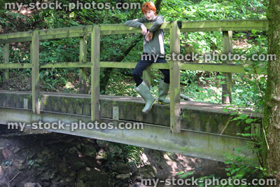 Stock image of teenage boy sitting on wooden bridge railings over woodland stream