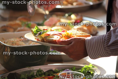 Stock image of buffet, person piling plate with party food, sandwich, Scotch egg