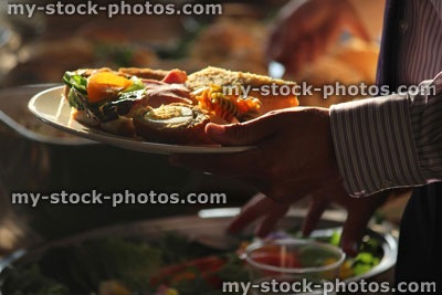 Stock image of buffet, person piling plate with party food, sandwich, Scotch egg