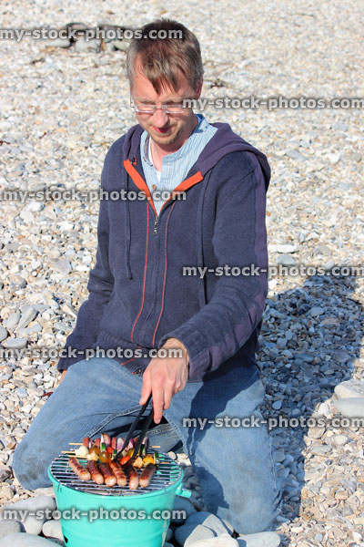 Stock image of man turning sausages on bucket barbecue at seaside