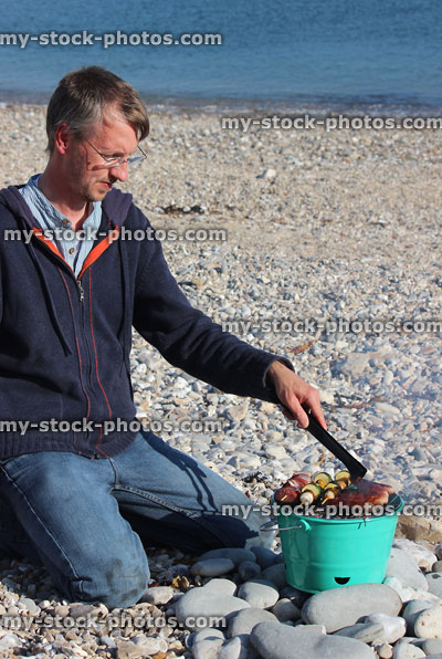 Stock image of man cooking sausages on beach barbecue using bucket