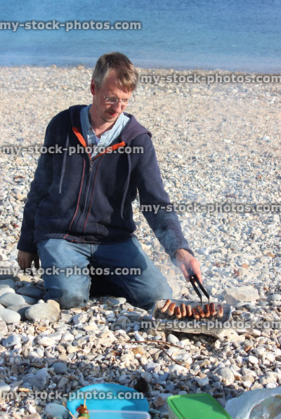 Stock image of man with charcoal barbecue, cooking sausages on pebble beach