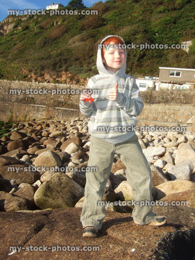 Stock image of young boy on large pebble beach, thumbs up, grey hoodie, eating chocolate bar
