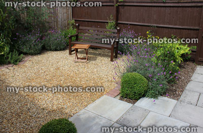 Stock image of wooden bench in gravel garden with flowers, shrubs, fence, table