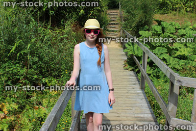 Stock image of girl standing in garden on sunny day, hat, sunglasses, summer dress, raised boardwalk