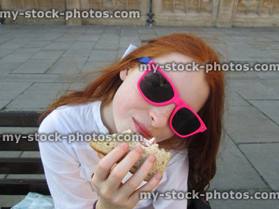Stock image of girl with long red hair, pink sunglasses, eating sandwich outside
