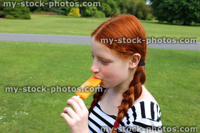 Stock image of girl eating frozen orange ice lolly in park