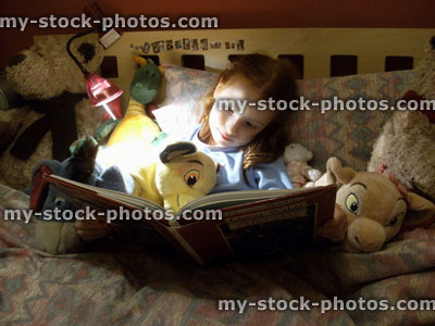 Stock image of young girl reading story book in bed, cuddly toys