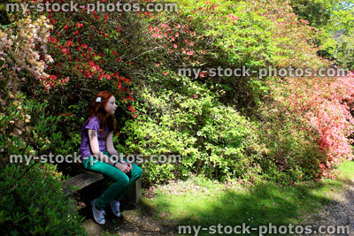 Stock image of girl sat on wooden garden bench by shrubs, trees, flowers