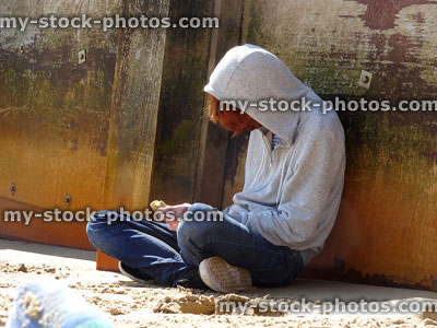 Stock image of hooded, teenage boy sat leaning against beach groyne