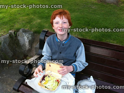 Stock image of young boy eating a chicken kebab takeaway outside on bench