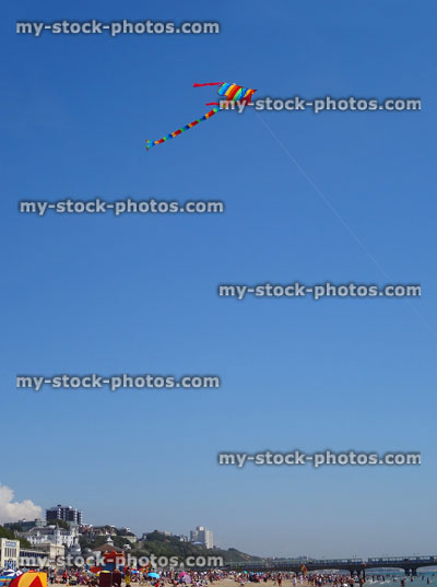 Stock image of stripy rainbow kite flying above seaside beach, blue sky
