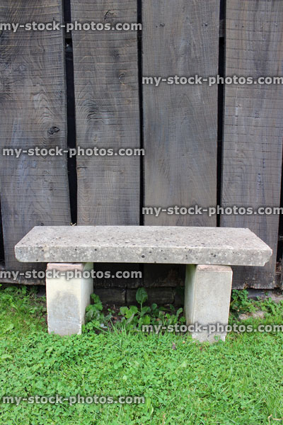 Stock image of concrete stone garden bench on green lawn, wooden fence slats