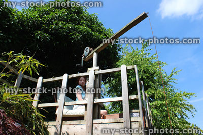 Stock image of girl sitting on treehouse decking platform, wooden crane toy, picket fence