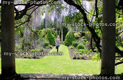 Stock image of girl running on garden lawn towards pergola of wisteria flowers